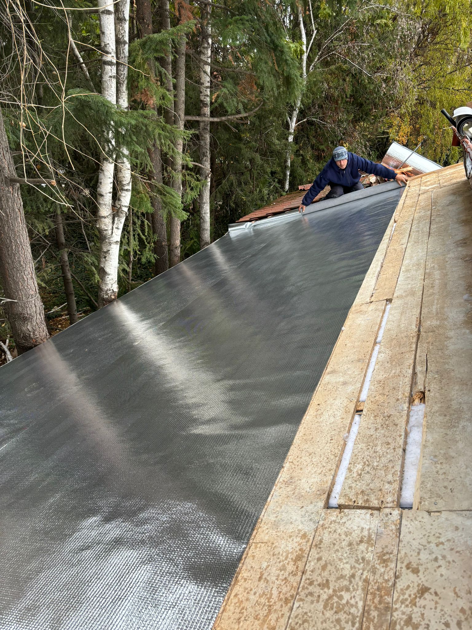 Derularea foliei anticondens Isoflect Roof Eco pe astereala de lemn a acoperișului — montaj în desfășurare, cu fâșia reflectivă întinsă dinspre coamă